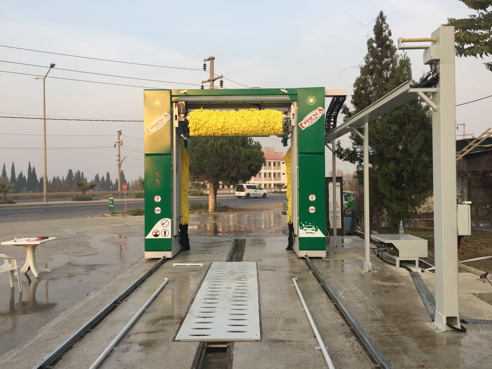 Green rollover car wash with a yellow top brush on floor rails at an outdoor wash bay, trees and a road behind